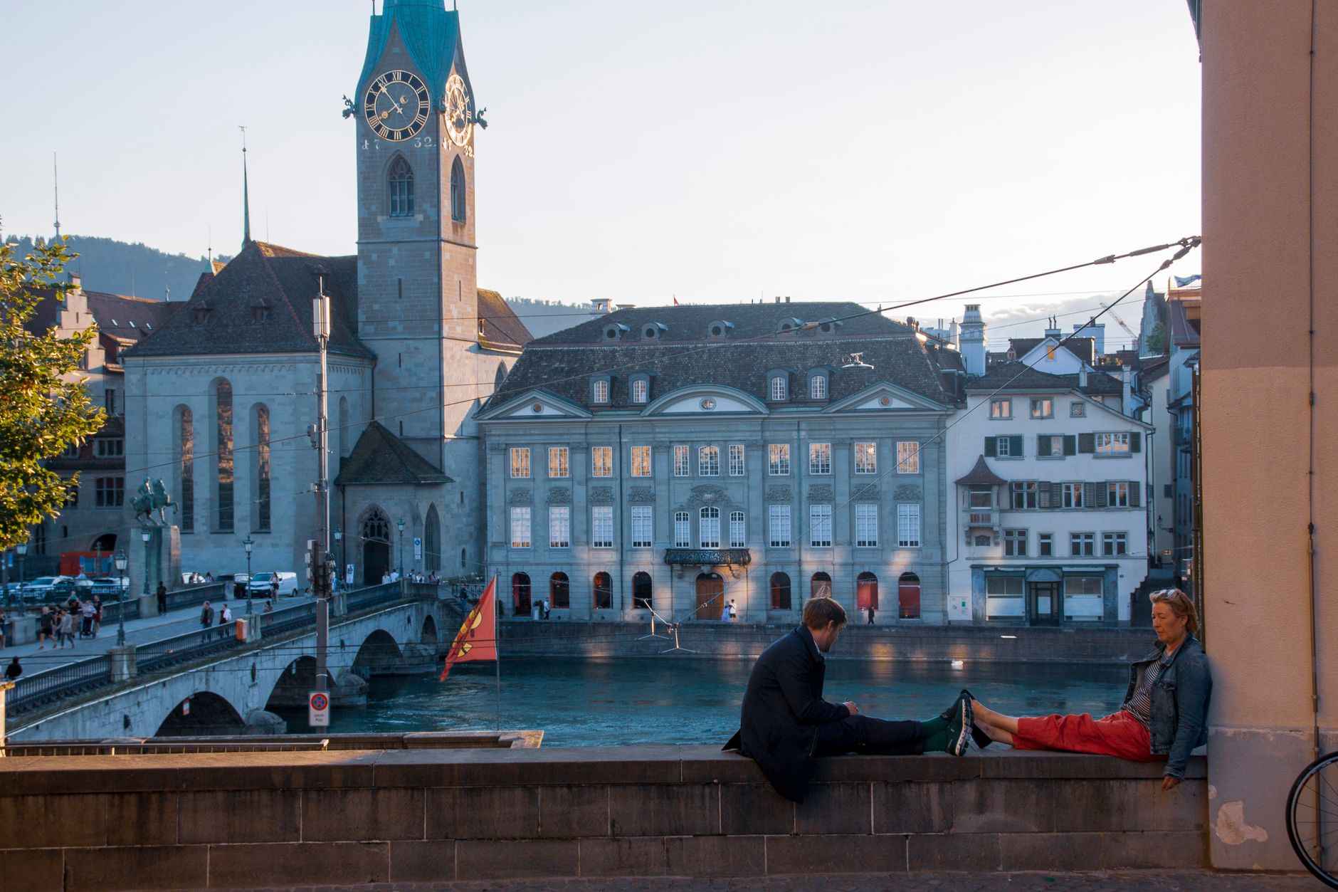 Two people relaxing by the river in Zurich's old town with scenic architecture and a historic clock tower.