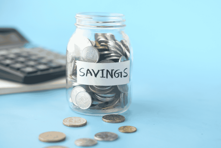 Glass jar filled with coins labeled savings next to a calculator on a blue background