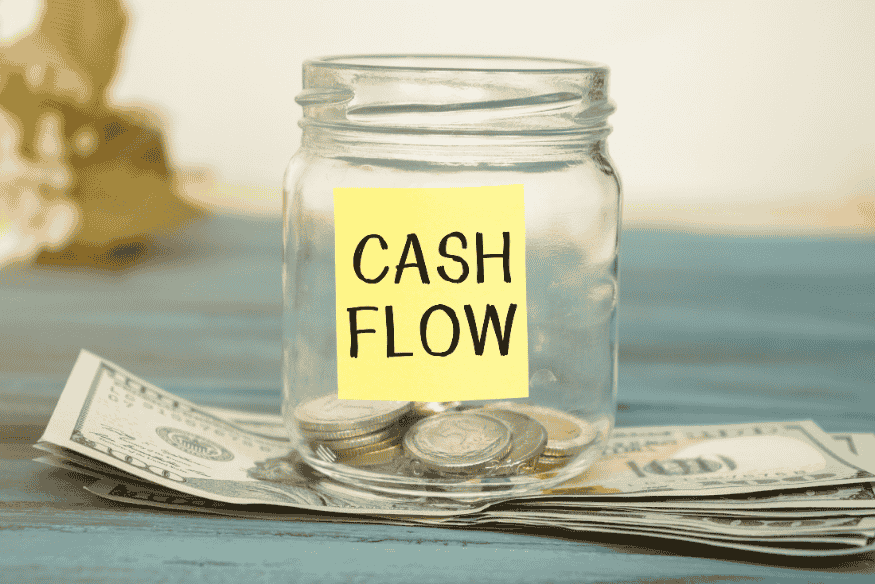 Glass jar labeled “cash flow” filled with coins and dollar bills on a wooden surface.