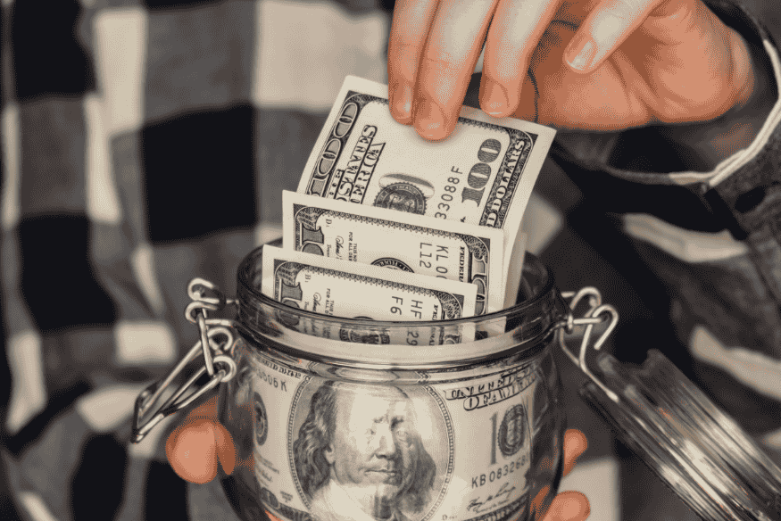 Person placing dollar bills into a savings jar to build financial stability from side income.