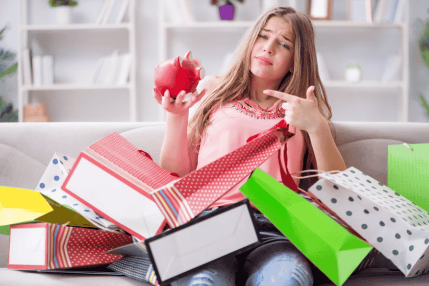 Young woman surrounded by colorful shopping bags looking at her piggy bank, illustrating impulse buying and the psychology behind overspending.